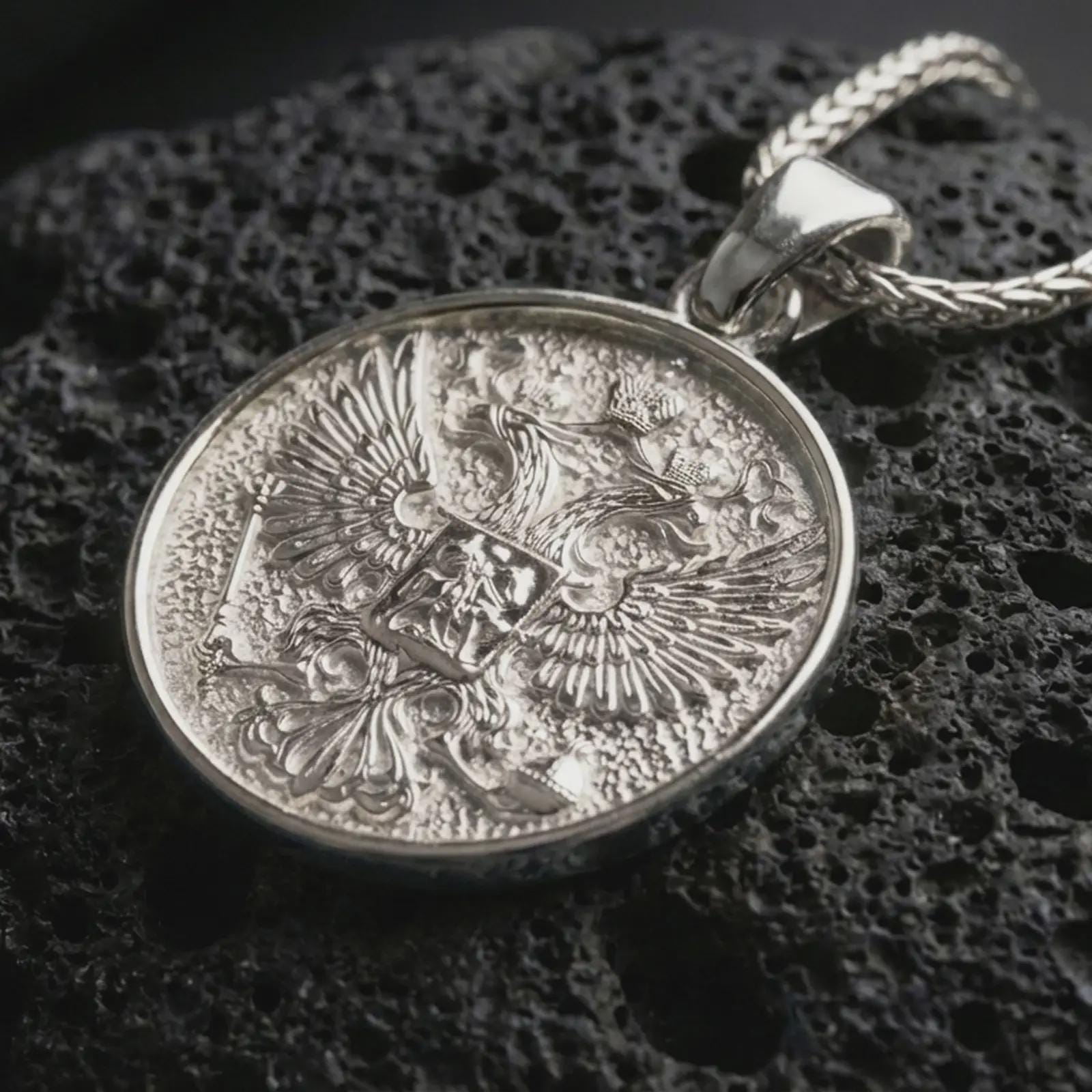 Polished silver double headed eagle pendant necklace displayed on dark volcanic rock, shown in a premium close-up image that highlights the imperial bird emblem, crown detail, and ornate relief texture.