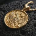 Load image into Gallery viewer, Gold plated double headed eagle pendant necklace photographed on porous black volcanic stone, emphasizing the historic heraldic symbol and round medallion form in a luxury jewelry composition.
