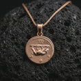 Load image into Gallery viewer, Rose gold plated Emperor Augustus coin pendant necklace presented on textured volcanic stone, showcasing the round Roman coin silhouette and refined emperor relief in a macro studio shot.
