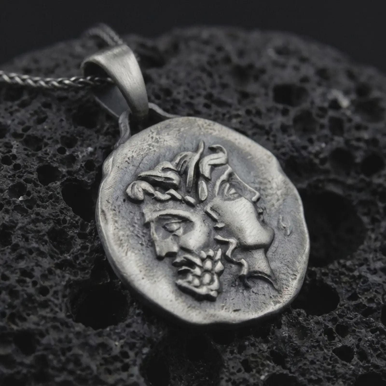 Oxidized Janus coin pendant necklace resting on black volcanic stone, featuring the ancient Roman two-faced god in a detailed antique-style relief and organic coin-shaped silhouette.