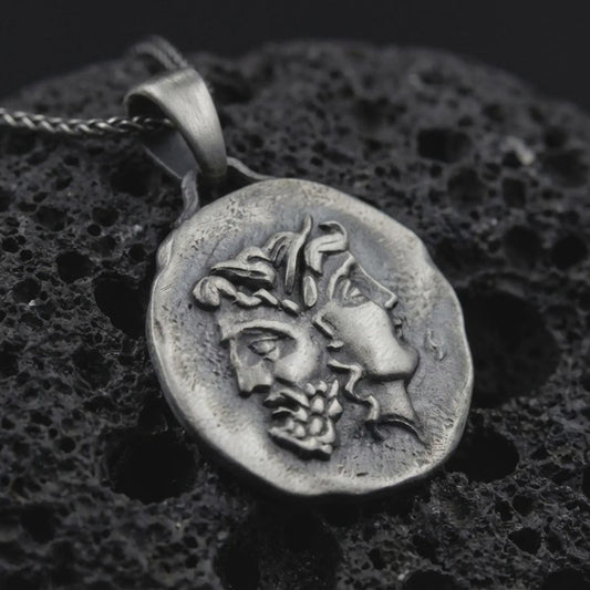Oxidized Janus coin pendant necklace resting on black volcanic stone, featuring the ancient Roman two-faced god in a detailed antique-style relief and organic coin-shaped silhouette.