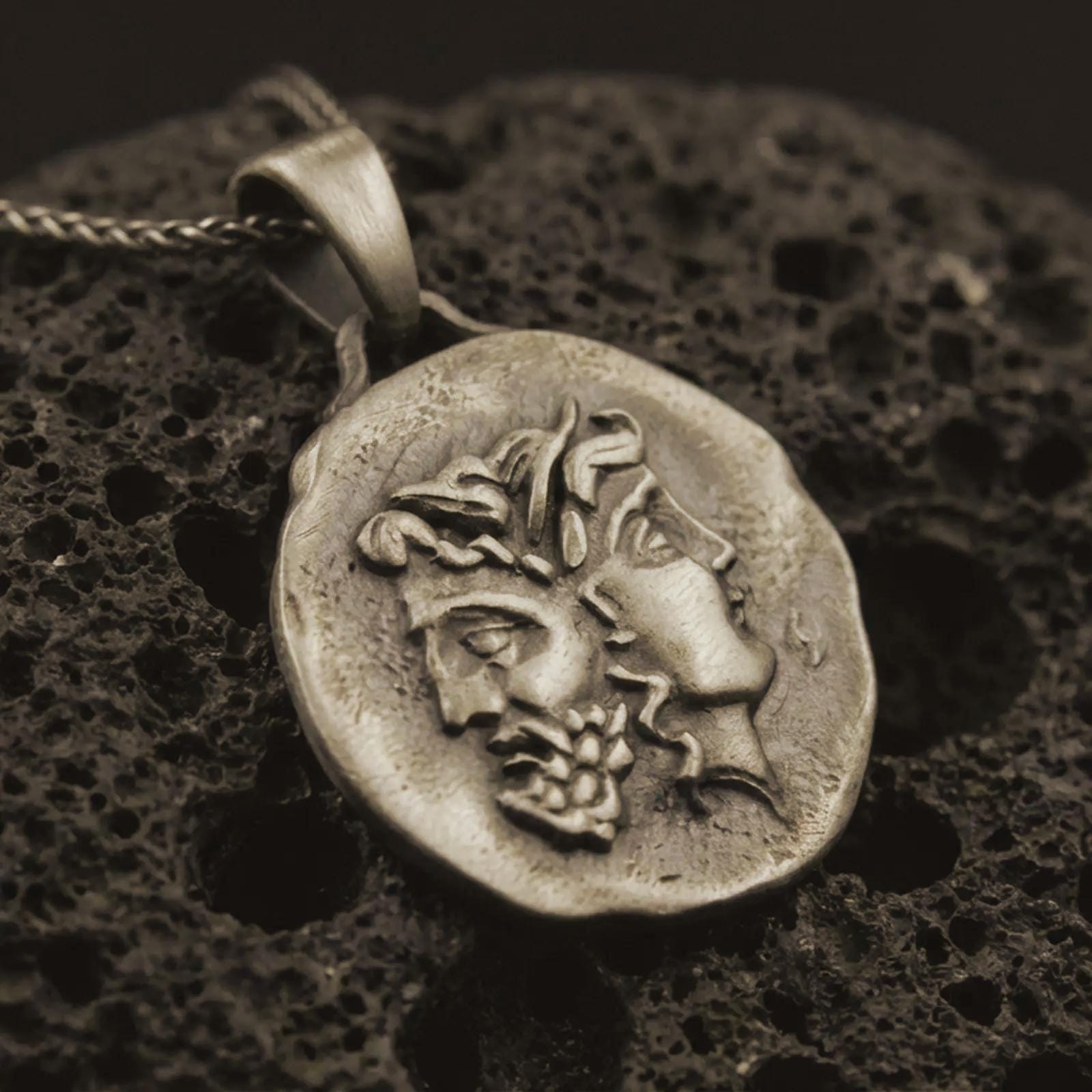 Bronze Janus coin pendant necklace resting on black volcanic stone, featuring the ancient Roman two-faced god in a detailed antique-style relief and organic coin-shaped silhouette.
