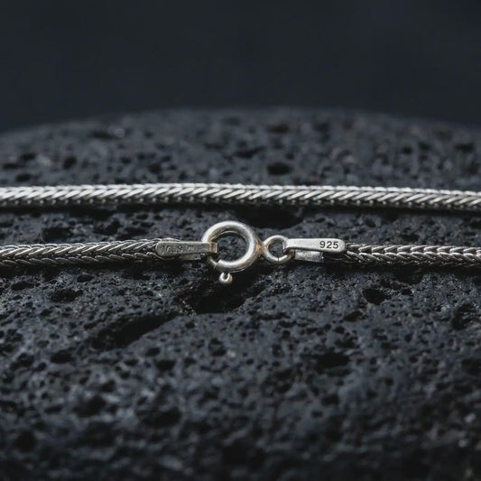 Polished silver Italian foxtail chain necklace on textured volcanic rock, centered macro jewelry photo with detailed wheat chain texture and bright silver highlights.