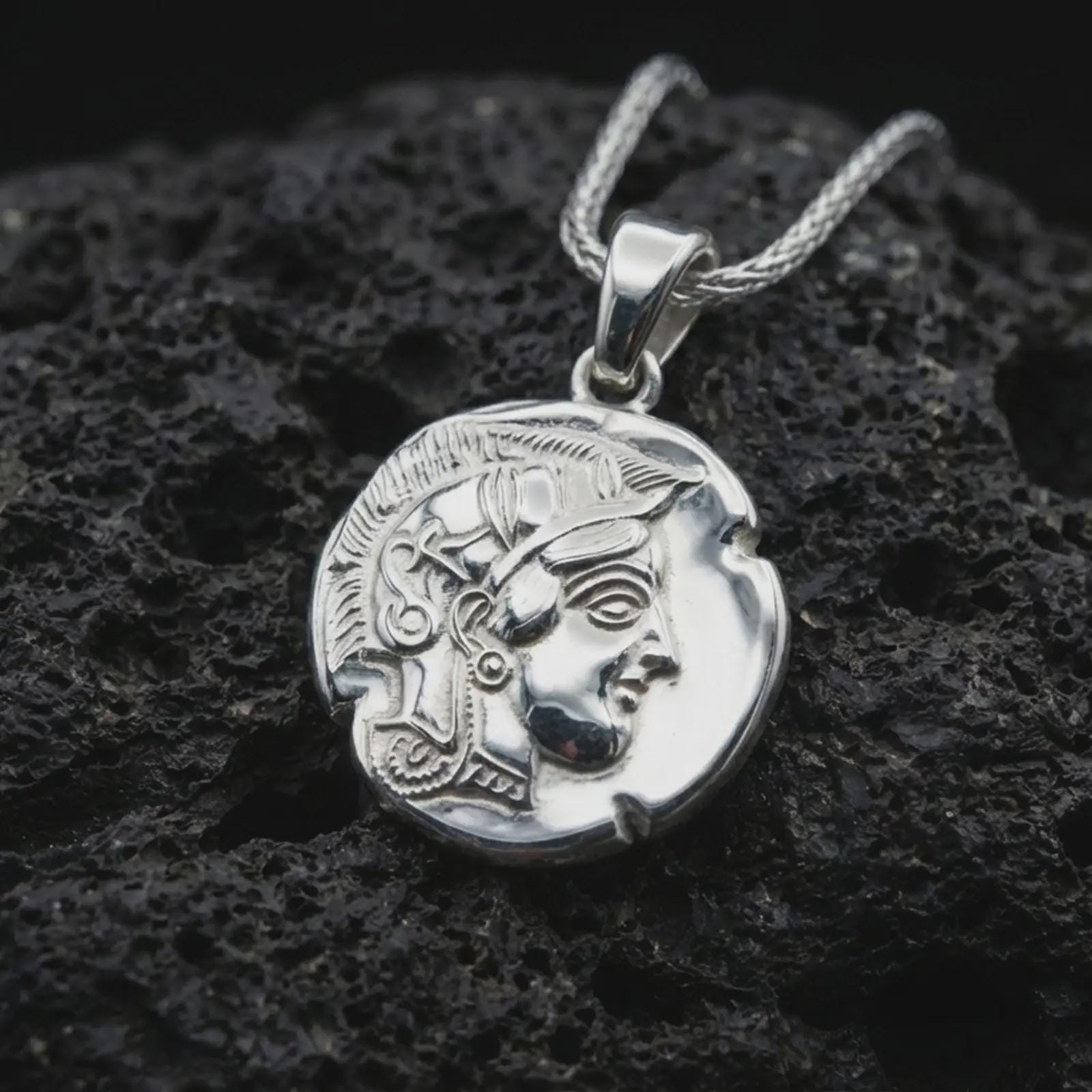 Polished silver Athena owl coin pendant necklace displayed on dark volcanic rock, shown in a premium close-up image that highlights the sacred owl motif and antique coin texture.