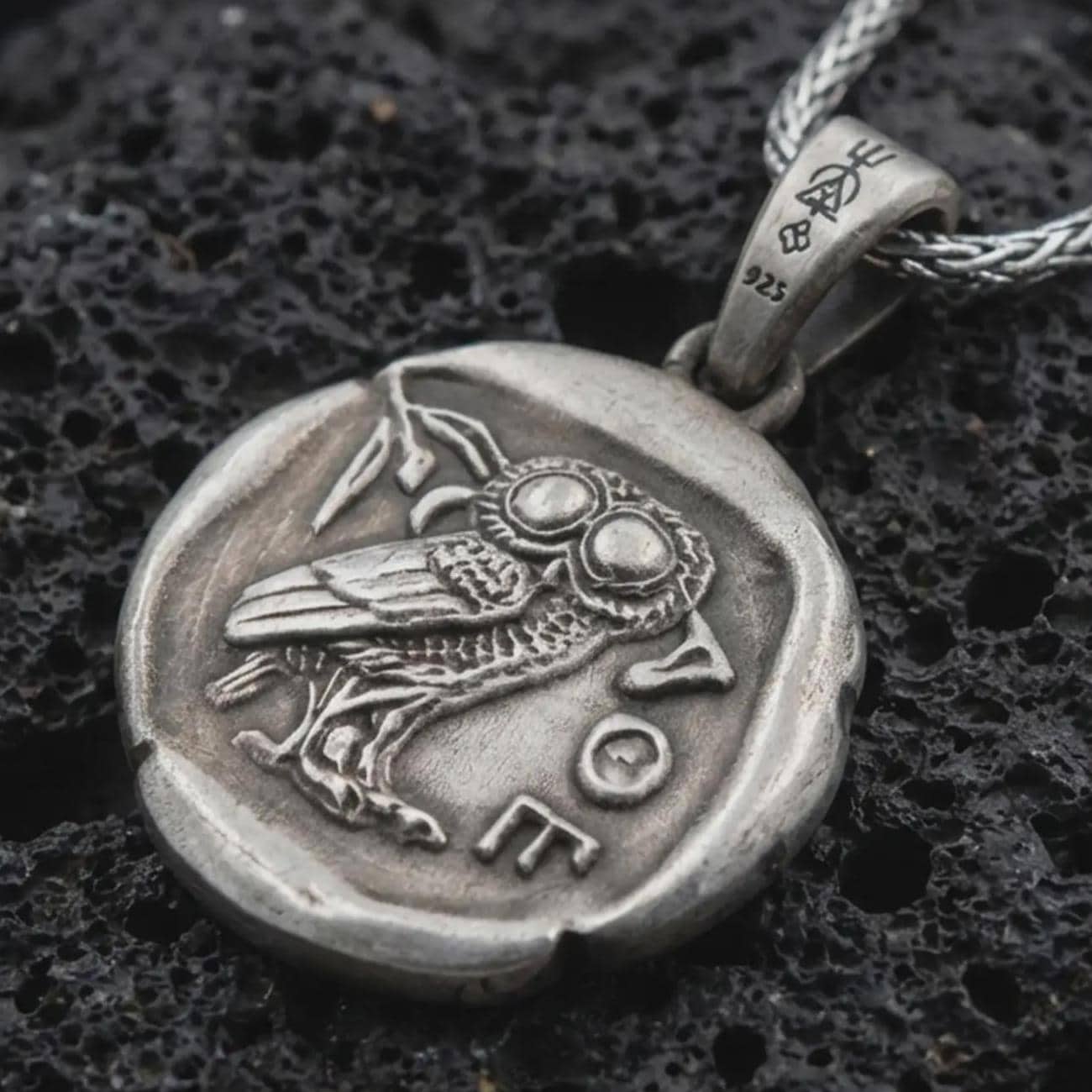 Oxidized Athena owl coin pendant necklace resting on black volcanic stone, featuring an ancient Greek coin-style owl relief with surrounding symbols in a detailed macro product photograph.