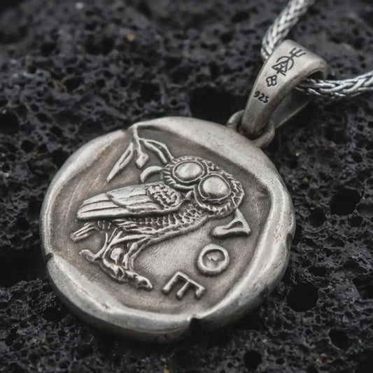 Oxidized Athena owl coin pendant necklace resting on black volcanic stone, featuring an ancient Greek coin-style owl relief with surrounding symbols in a detailed macro product photograph.