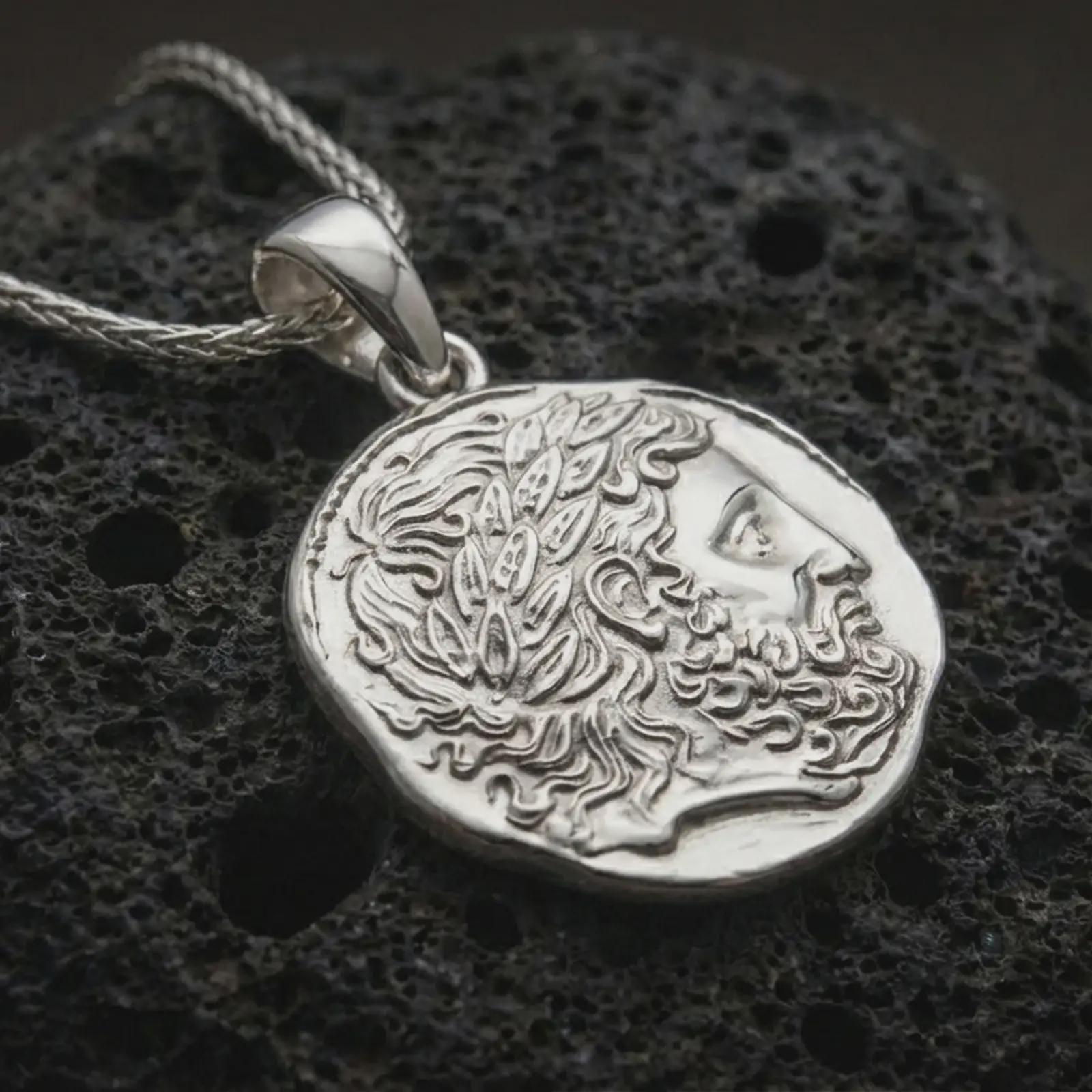Polished silver ancient Greek coin pendant necklace displayed on dark volcanic rock, shown in a premium close-up image that highlights the stylized male portrait and organic antique coin texture.