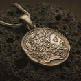 Load image into Gallery viewer, Bronze ancient Greek coin pendant necklace resting on black volcanic stone, featuring a bearded classical profile with flowing hair and laurel-like ornament in a detailed macro product photograph.
