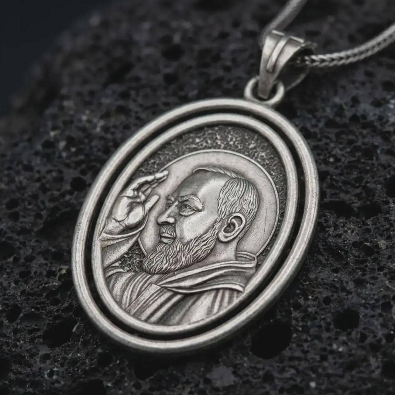 Oxidized Saint Padre Pio spinning pendant necklace resting on black volcanic stone, featuring an oval double-frame medal with a detailed portrait of Padre Pio raising his hand in blessing.