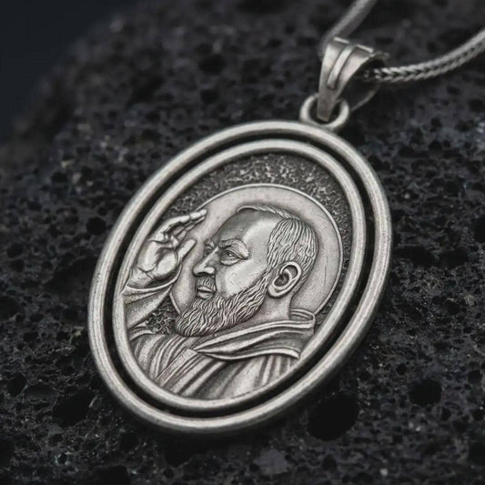 Oxidized Saint Padre Pio spinning pendant necklace resting on black volcanic stone, featuring an oval double-frame medal with a detailed portrait of Padre Pio raising his hand in blessing.