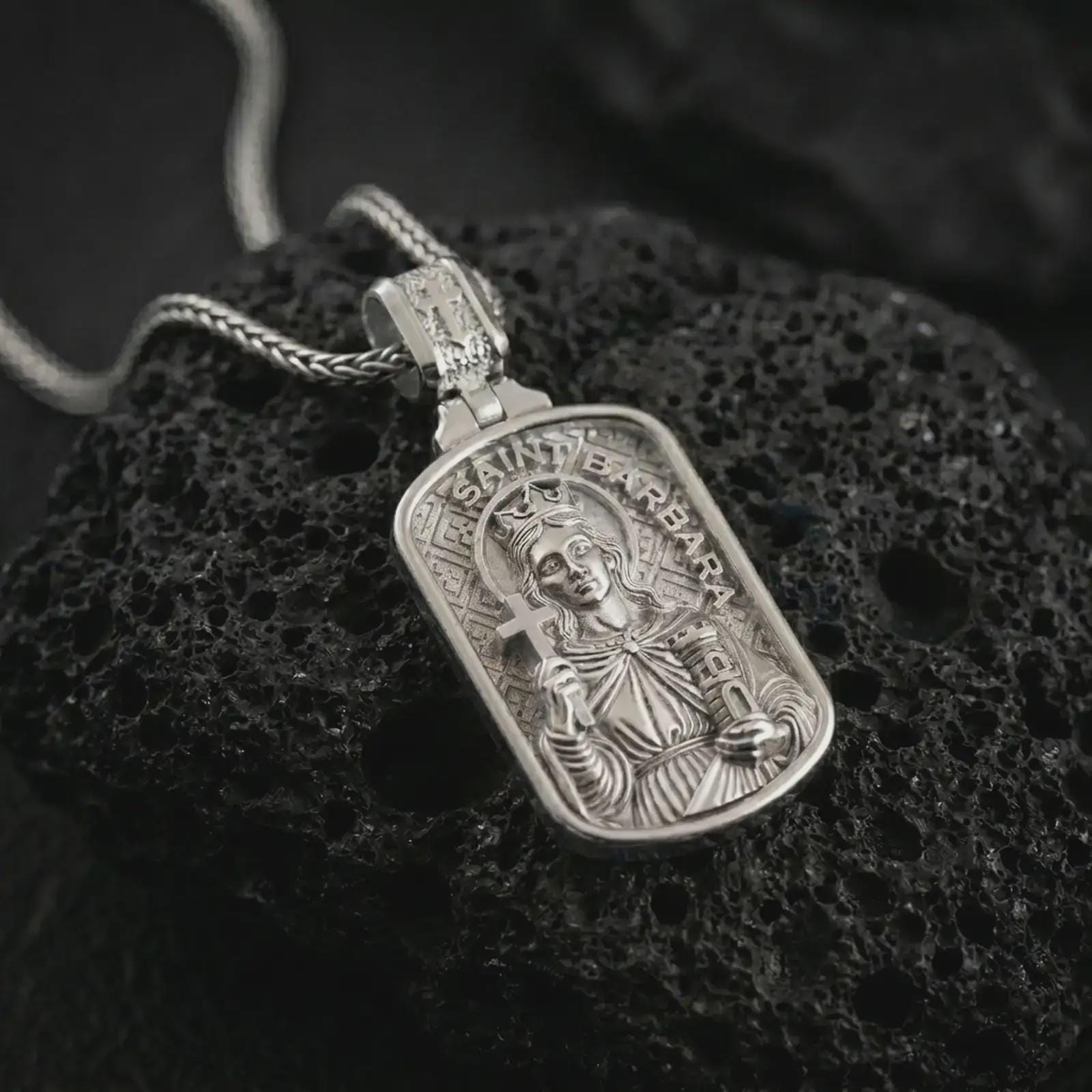 Polished silver Saint Barbara pendant necklace on textured volcanic rock, centered macro jewelry photo with detailed religious icon engraving and bright silver highlights.