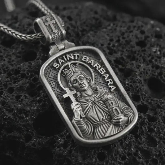 Oxidized Saint Barbara pendant necklace on textured volcanic rock, close-up studio shot highlighting the saint relief, cross, tower detail, and dark antique silver contrast.