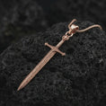 Load image into Gallery viewer, Rose gold plated medieval sword pendant necklace on textured volcanic rock, detailed macro product shot with soft warm reflections and gothic sword symbolism.

