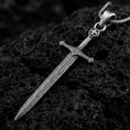 Load image into Gallery viewer, Oxidized medieval sword pendant necklace on textured volcanic rock, close-up studio shot highlighting the long blade, ornate hilt, cross detail, and antique silver contrast.
