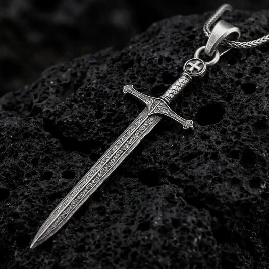Oxidized medieval sword pendant necklace on textured volcanic rock, close-up studio shot highlighting the long blade, ornate hilt, cross detail, and antique silver contrast.