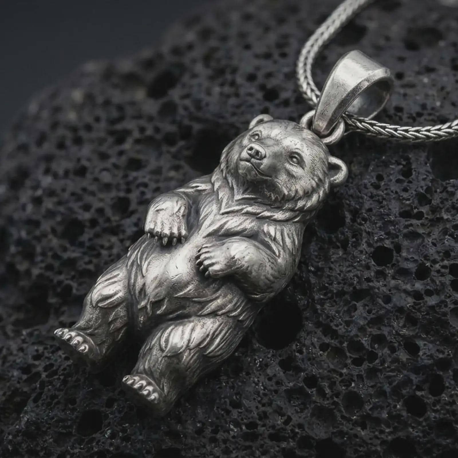 Oxidized baby bear pendant necklace on textured volcanic rock, close-up studio shot highlighting the cute bear cub charm, rounded paws, detailed fur, and antique silver contrast.