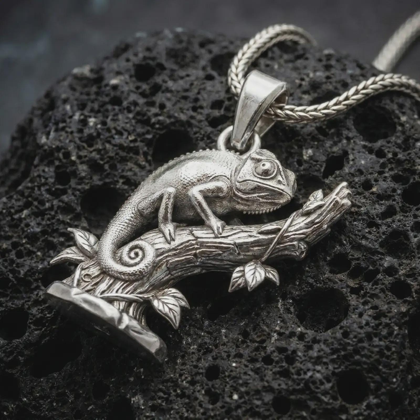 Polished silver chameleon pendant necklace on textured volcanic rock, centered macro jewelry photo with detailed reptile texture, branch relief, and bright silver highlights.