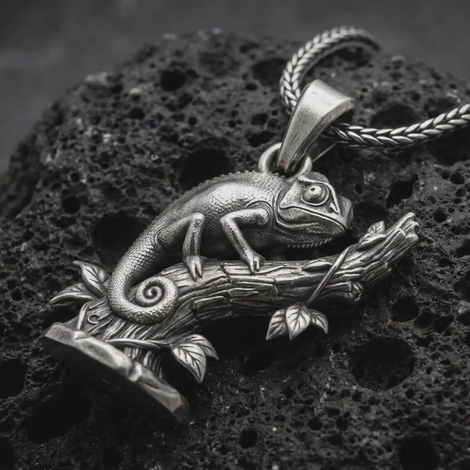 Oxidized chameleon pendant necklace on textured volcanic rock, close-up studio shot highlighting the lizard body, curled tail, branch detail, leaves, and antique silver contrast.