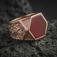 Load image into Gallery viewer, A red onyx signet ring in a rose gold plated finish resting on porous volcanic rock in a premium macro studio shot.
