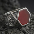 Load image into Gallery viewer, A red onyx signet ring in an oxidized finish resting on porous volcanic rock in a premium macro studio shot.
