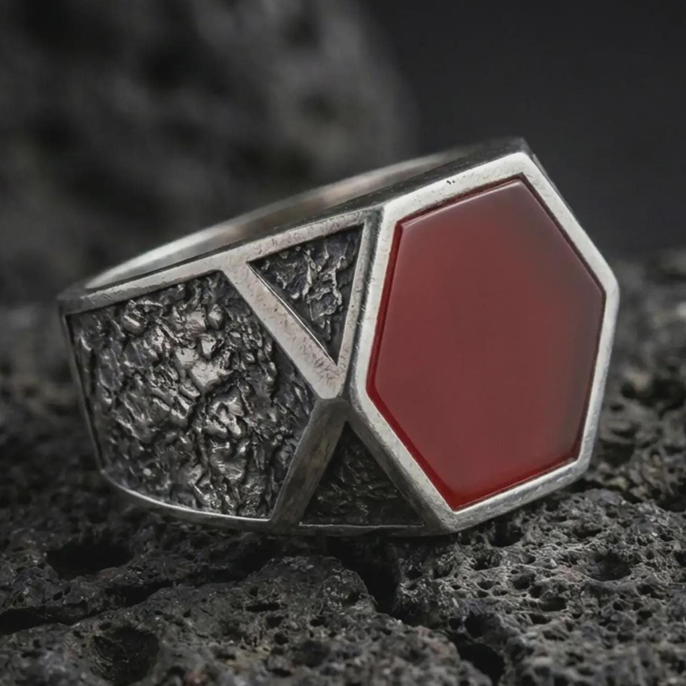 A red onyx signet ring in an oxidized finish resting on porous volcanic rock in a premium macro studio shot.