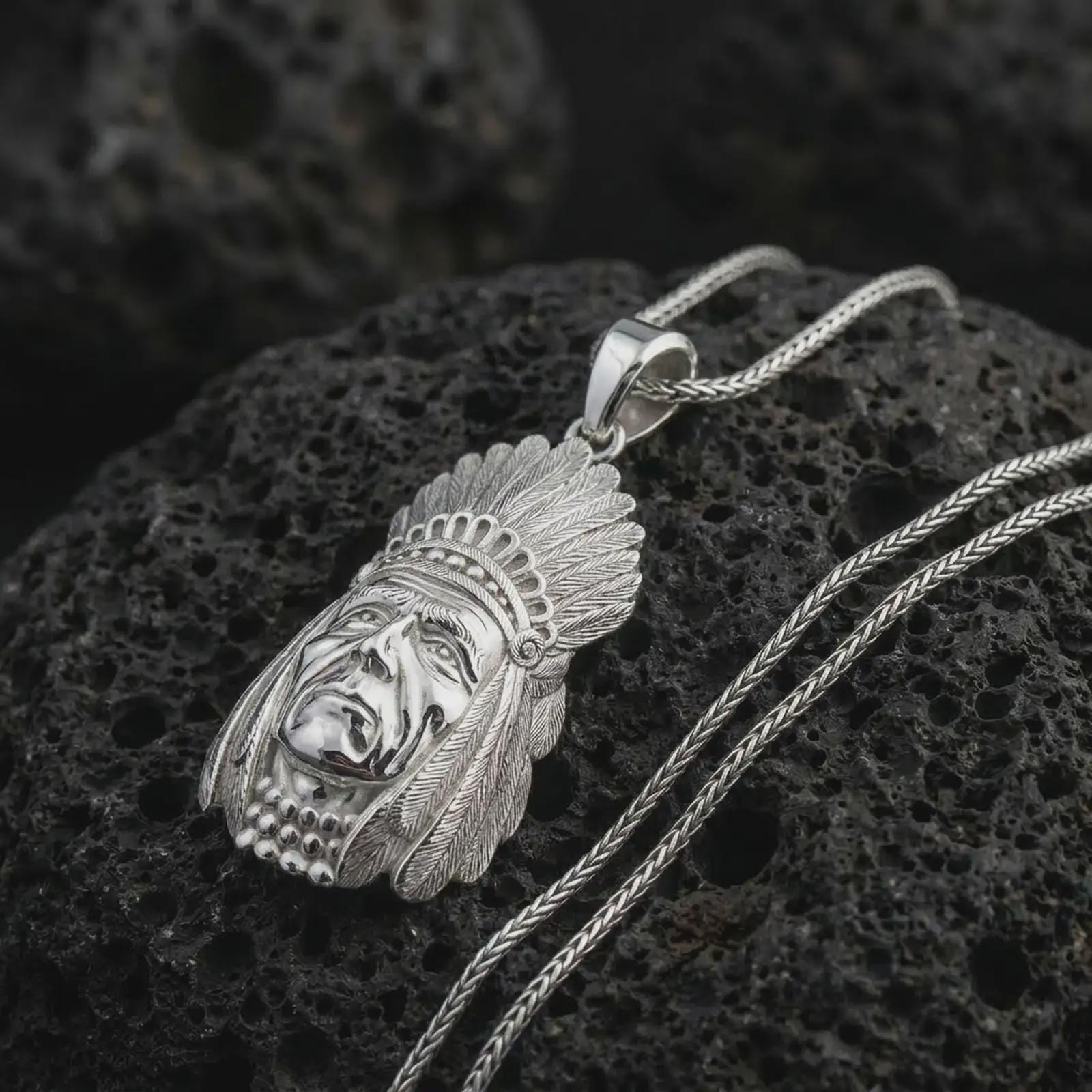 Polished silver feather headdress chief pendant necklace on textured volcanic rock, centered macro jewelry photo with detailed portrait relief and bright silver highlights.
