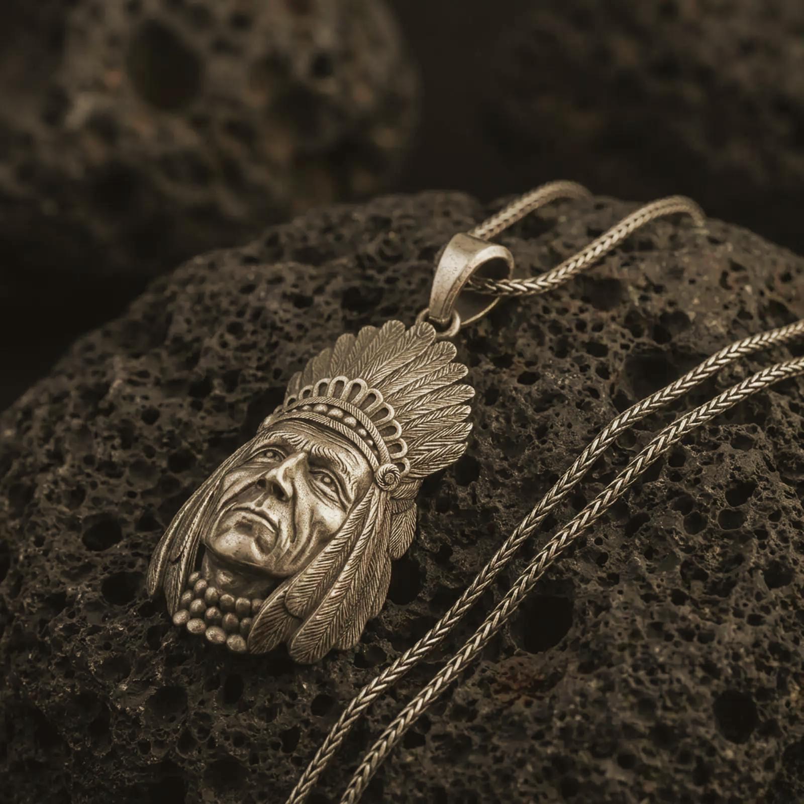 Bronze feather headdress chief pendant necklace on textured volcanic rock, close-up studio shot highlighting the sculpted face, feather details, beaded necklace, and antique silver contrast.