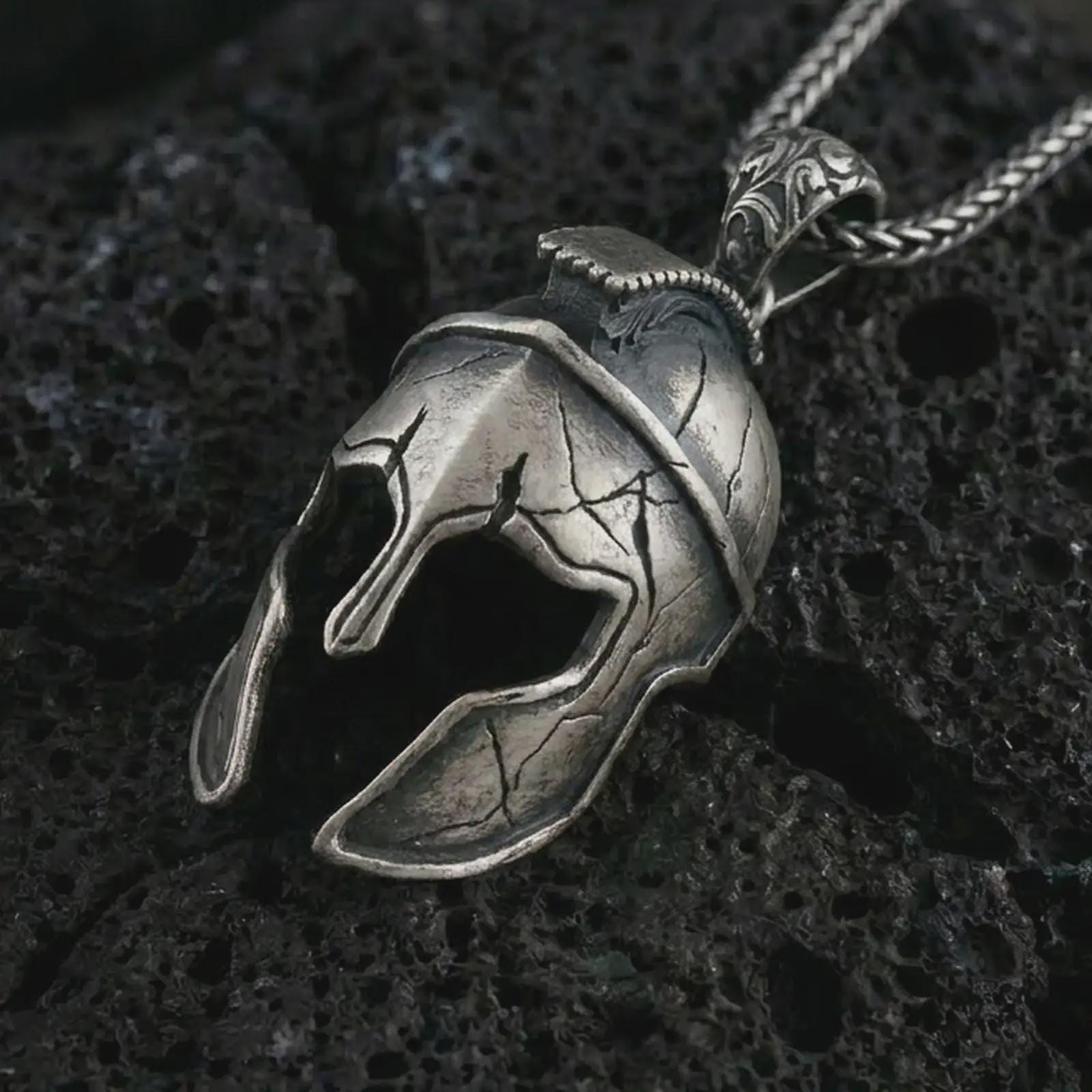 Oxidized Spartan helmet pendant necklace on textured volcanic rock, close-up studio shot highlighting the cracked warrior helmet, open face guard, battle-worn texture, and antique silver contrast.