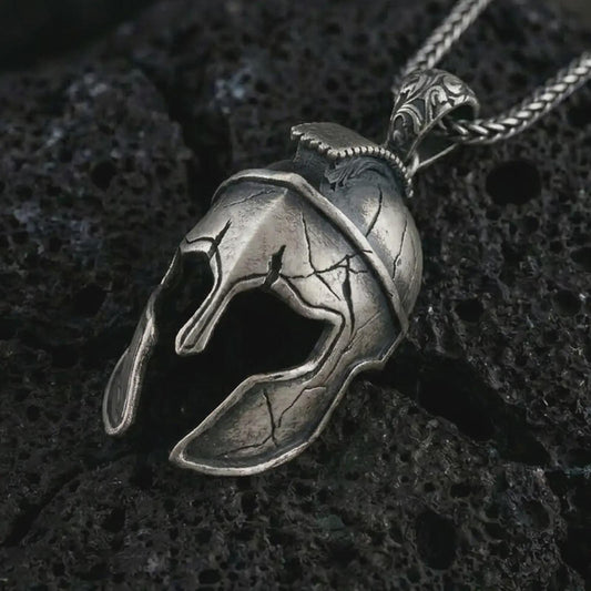 Oxidized Spartan helmet pendant necklace on textured volcanic rock, close-up studio shot highlighting the cracked warrior helmet, open face guard, battle-worn texture, and antique silver contrast.