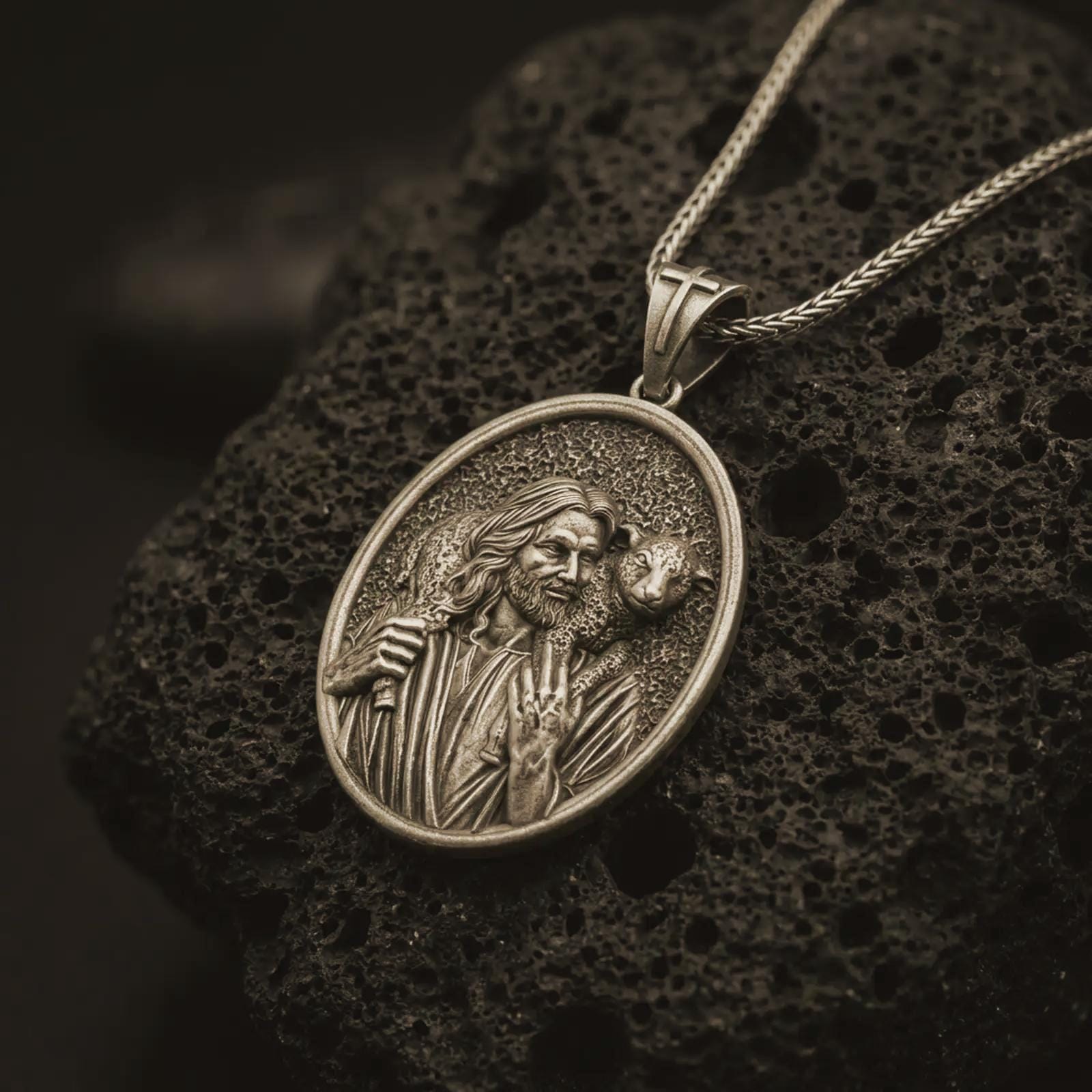 Bronze Good Shepherd Jesus pendant necklace on textured volcanic rock, close-up studio shot highlighting the shepherd relief, lamb detail, and antique silver contrast.