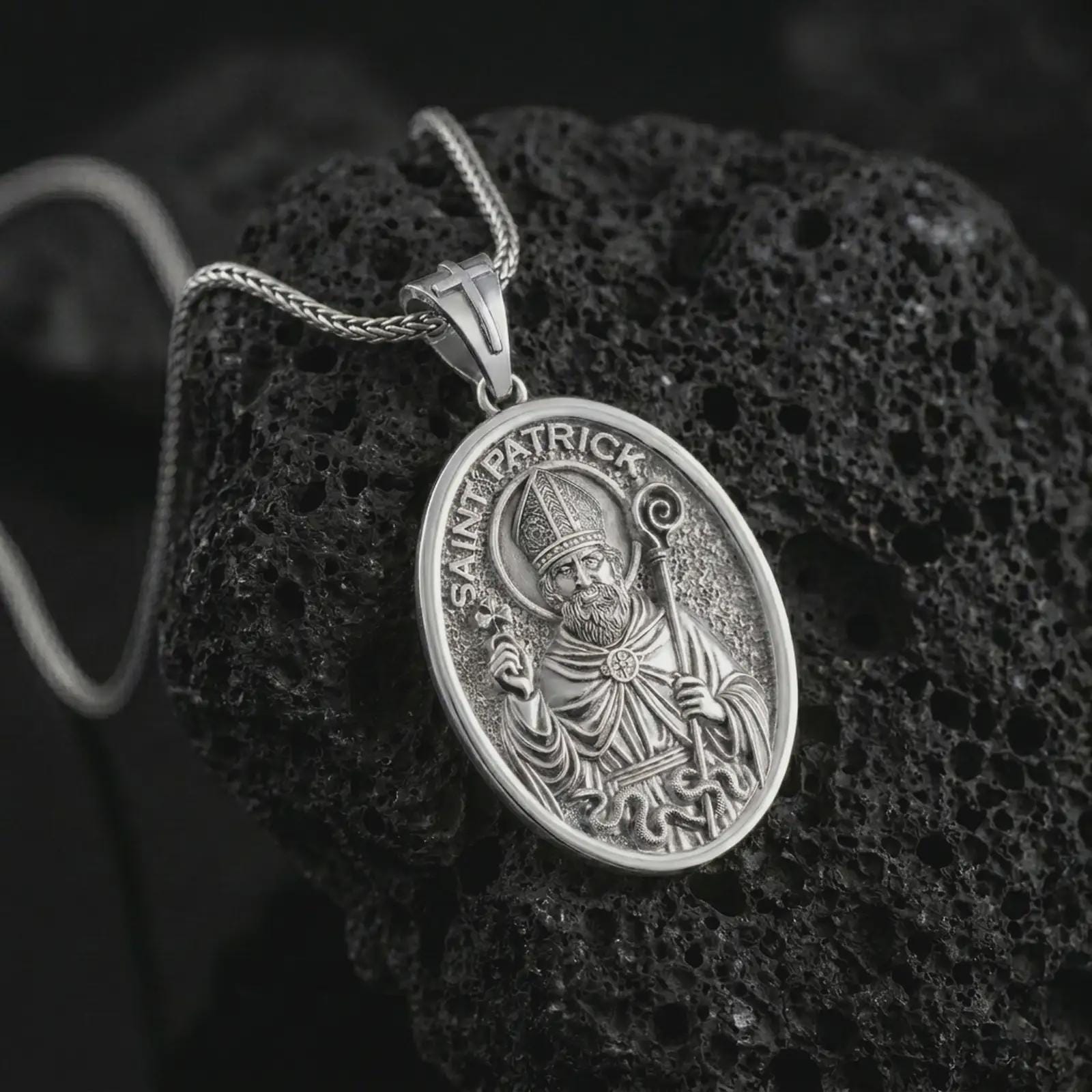 Polished silver Saint Patrick pendant necklace on textured volcanic rock, centered macro jewelry photo with detailed Irish patron saint engraving and bright silver highlights.