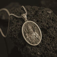 Load image into Gallery viewer, Bronze Saint Patrick pendant necklace on textured volcanic rock, close-up studio shot highlighting the bishop relief, shamrock, staff, and antique silver contrast.
