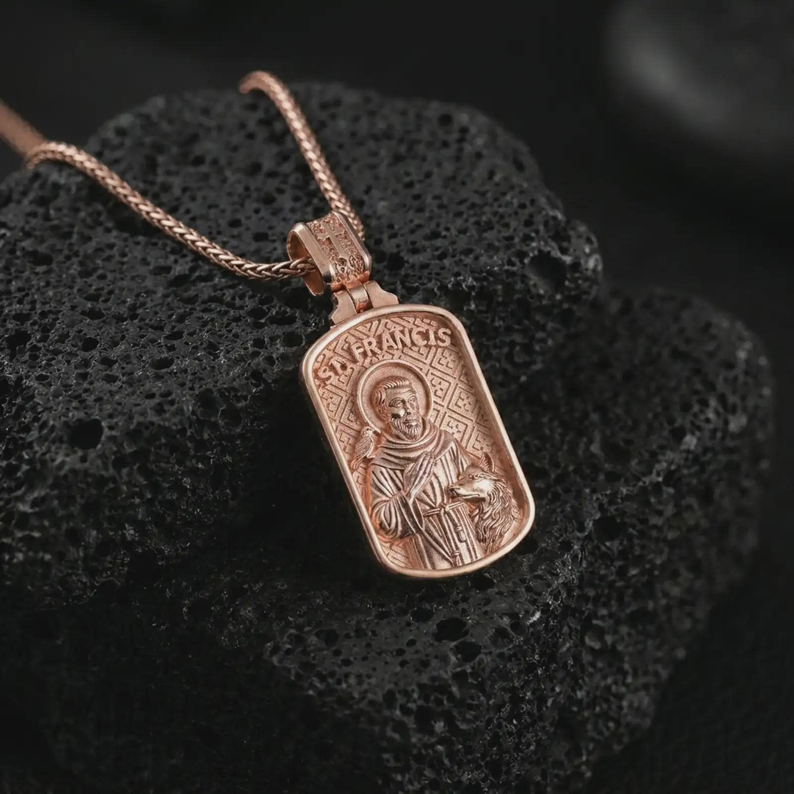 Rose gold plated Saint Francis pendant necklace on textured volcanic rock, detailed macro product shot with soft warm reflections and devotional Saint Francis design.