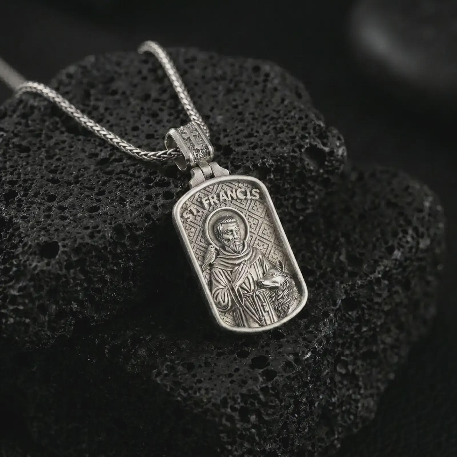 Polished silver Saint Francis pendant necklace on textured volcanic rock, centered macro jewelry photo with detailed religious engraving and bright silver highlights.