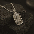 Load image into Gallery viewer, Bronze Saint Francis pendant necklace on textured volcanic rock, close-up studio shot highlighting the saint relief, bird, wolf detail, and antique silver contrast.
