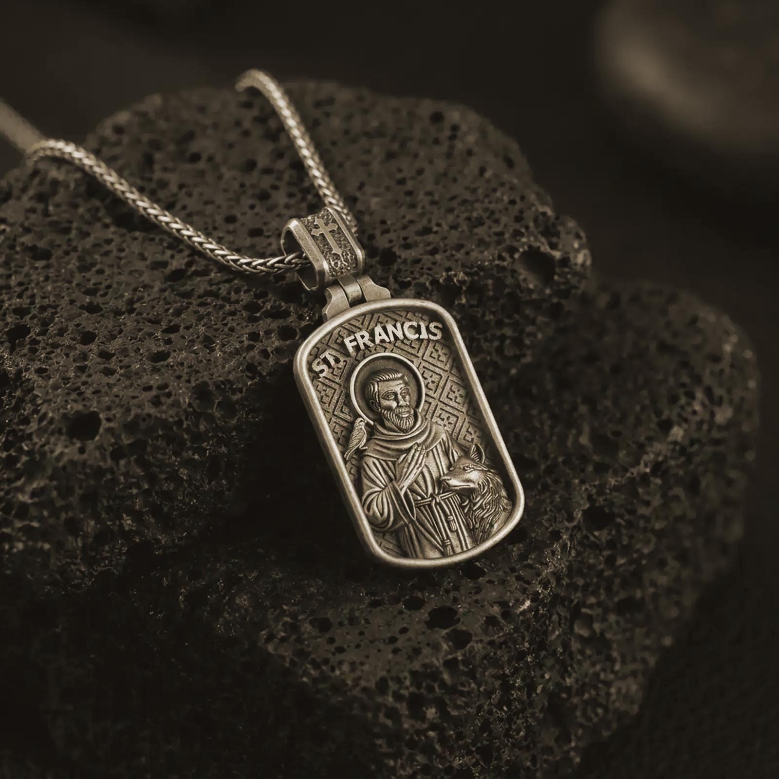 Bronze Saint Francis pendant necklace on textured volcanic rock, close-up studio shot highlighting the saint relief, bird, wolf detail, and antique silver contrast.
