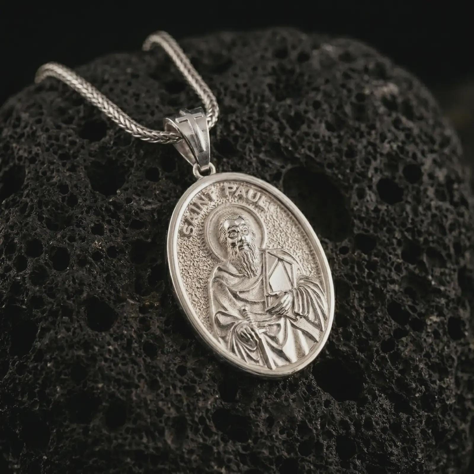 Polished silver Saint Paul pendant necklace on textured volcanic rock, centered macro jewelry photo with detailed Catholic medal engraving and bright silver highlights.