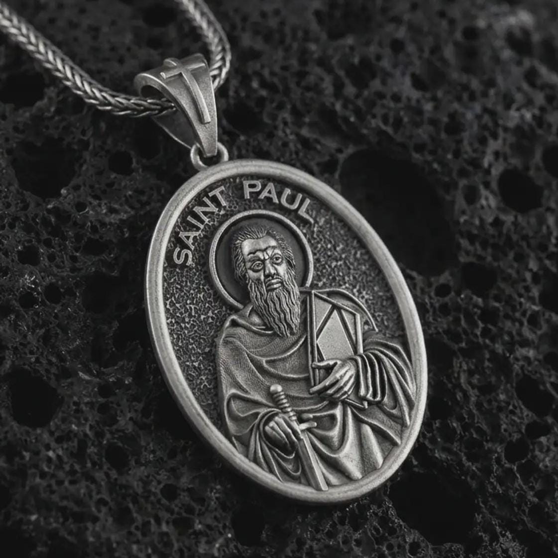 Oxidized Saint Paul pendant necklace on textured volcanic rock, close-up studio shot highlighting the apostle relief, sword, book detail, and antique silver contrast.