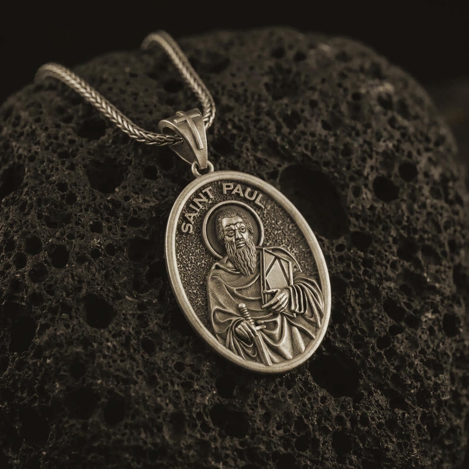 Bronze Saint Paul pendant necklace on textured volcanic rock, close-up studio shot highlighting the apostle relief, sword, book detail, and antique silver contrast.