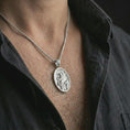 Load image into Gallery viewer, Polished silver Saint Francis pendant necklace worn on the chest, masculine lifestyle shot highlighting the saint relief, bird, wolf detail, and bright silver finish.
