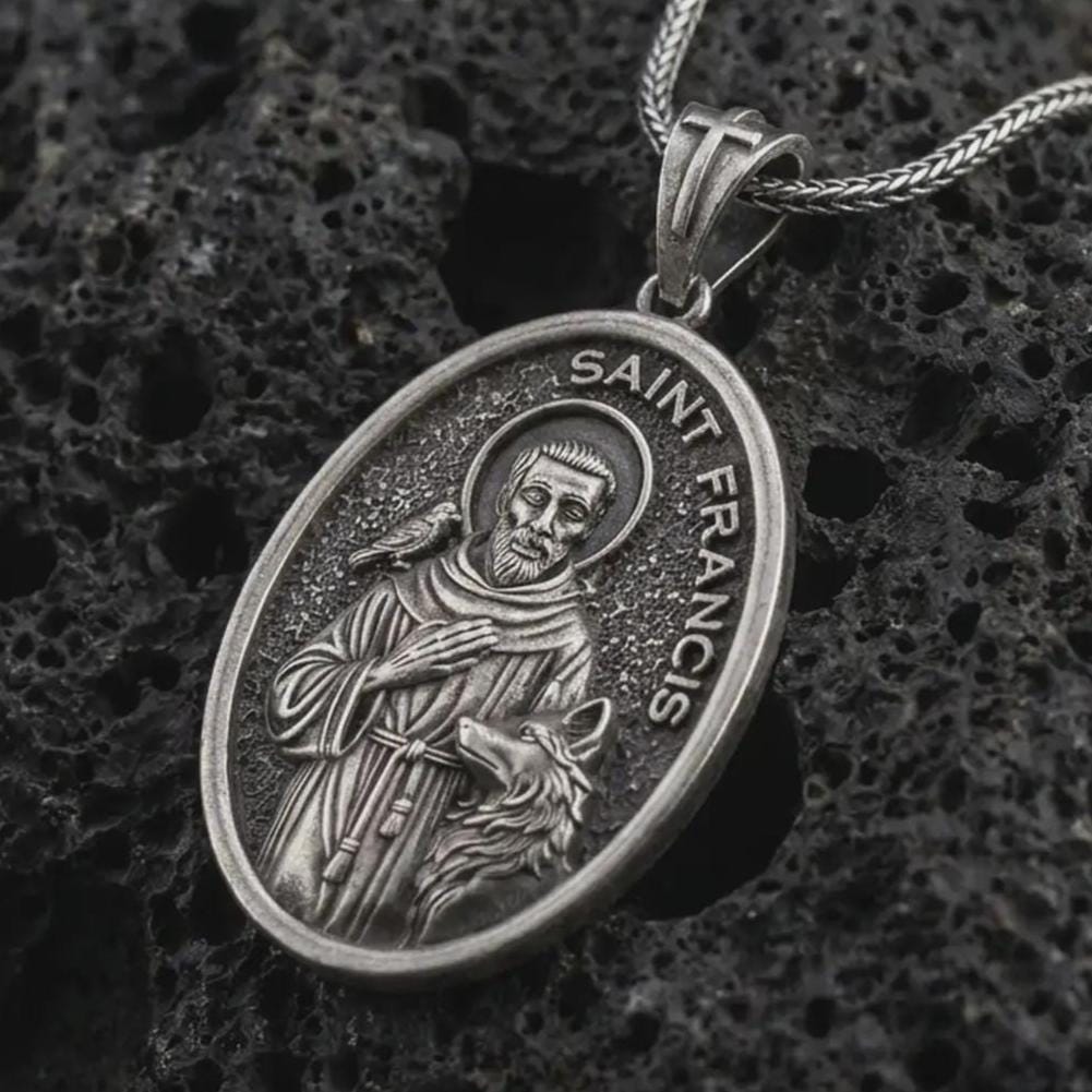 Oxidized Saint Francis pendant necklace on textured volcanic rock, close-up studio shot highlighting the saint relief, bird, wolf detail, and antique silver contrast.