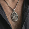Load image into Gallery viewer, Oxidized Saint Andrew pendant necklace worn on the chest, close-up lifestyle jewelry photo showing the religious medallion on a spiga chain with dark clothing.

