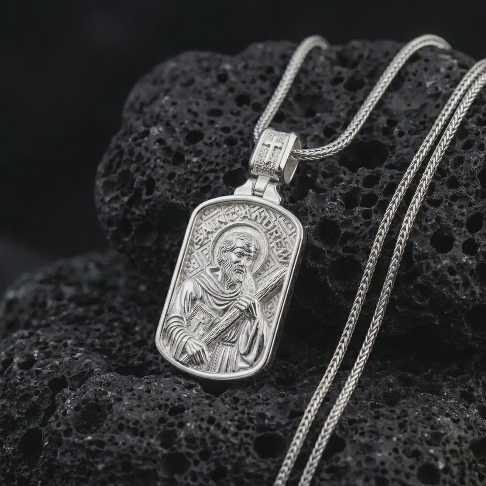 Polished silver Saint Andrew dog tag pendant necklace on textured volcanic rock, centered macro jewelry photo with detailed Catholic medal engraving and bright silver highlights.