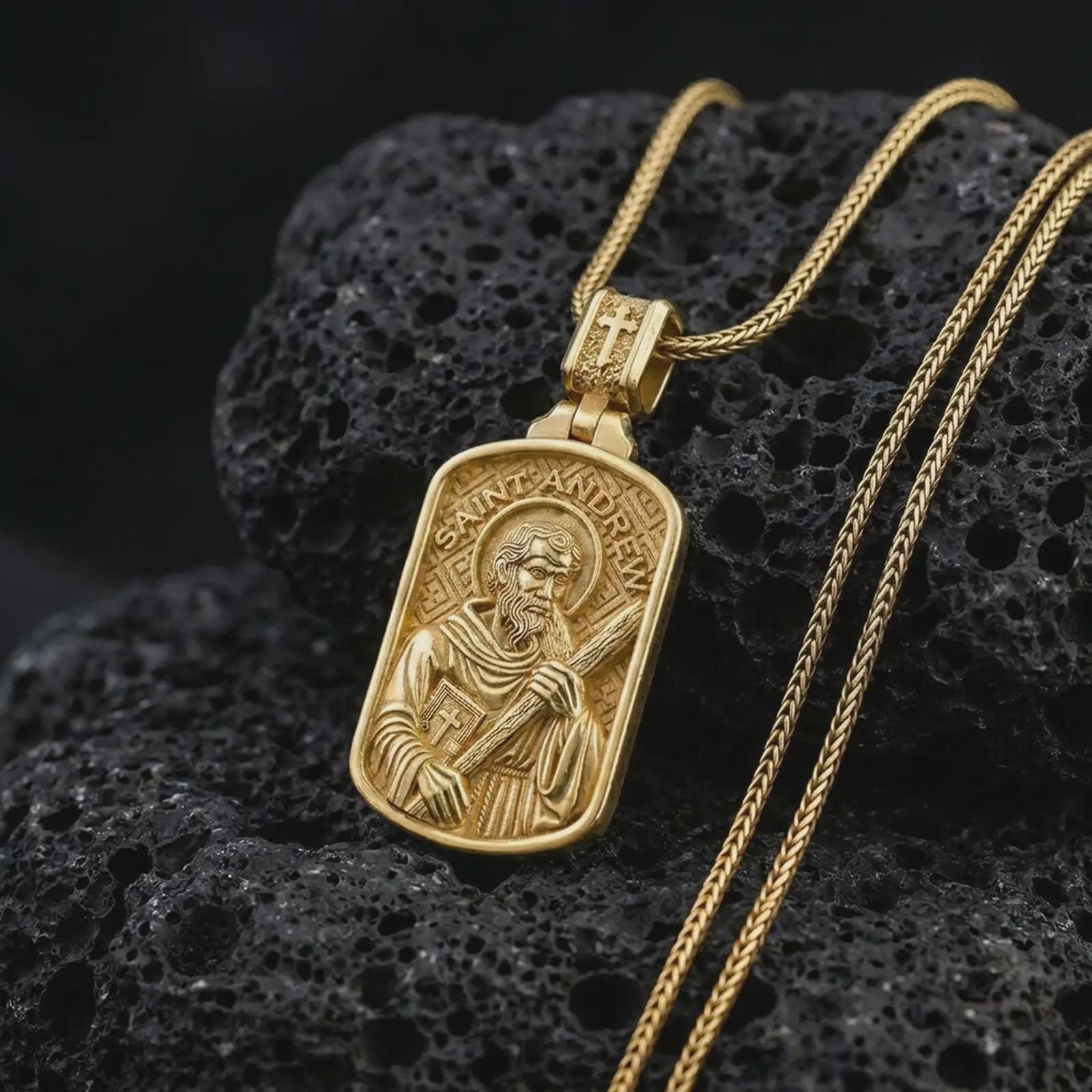 Gold plated Saint Andrew dog tag pendant necklace on textured volcanic rock, premium close-up studio composition emphasizing the apostle figure, cross, and sacred book symbolism.