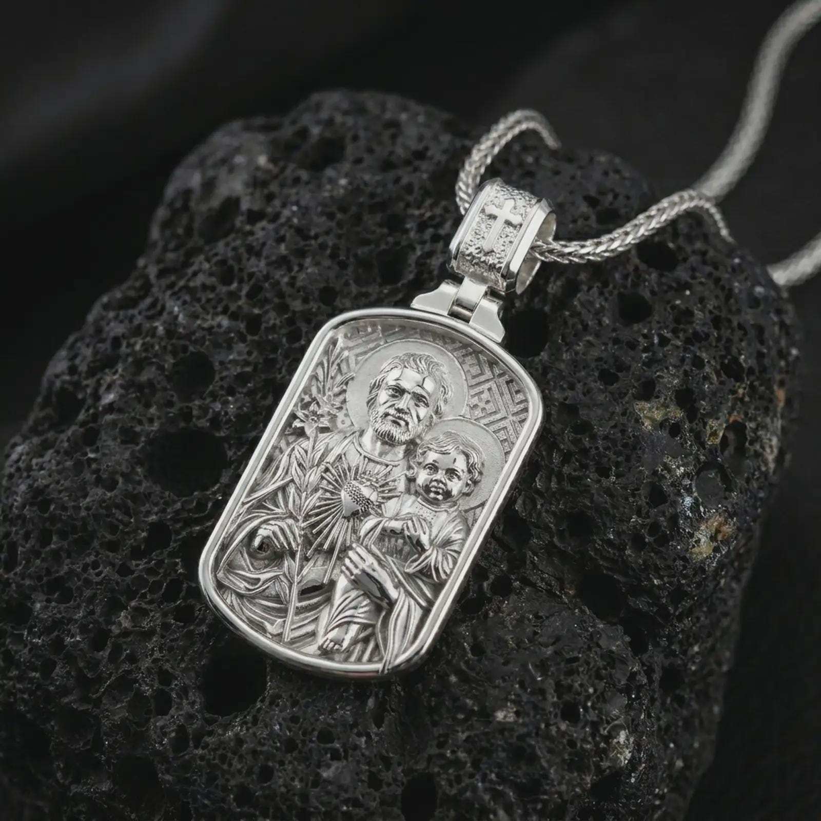 Polished silver Saint Joseph and Jesus pendant necklace on textured volcanic rock, centered macro jewelry photo with detailed Catholic medal engraving and bright silver highlights.