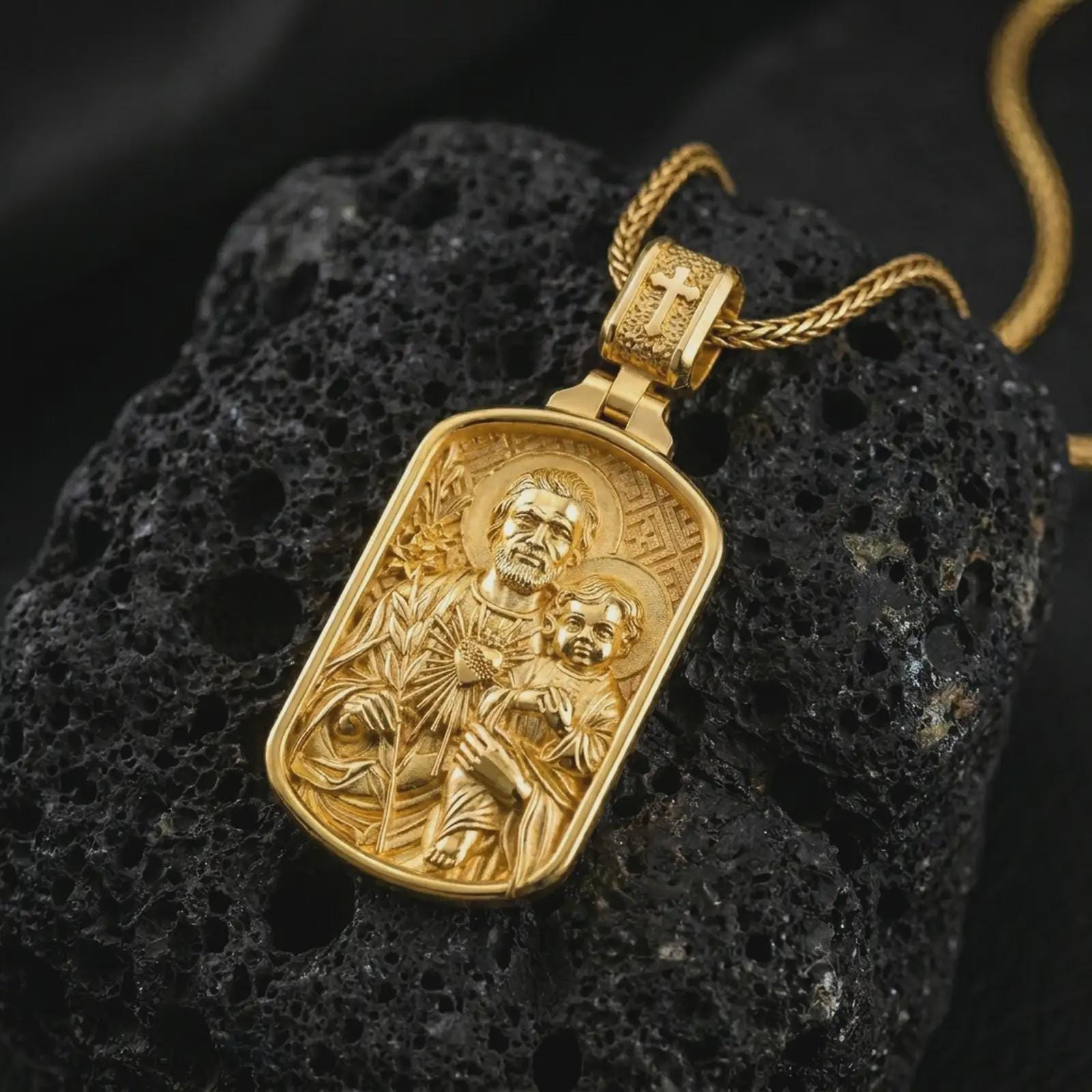 Gold plated Saint Joseph and Jesus pendant necklace on textured volcanic rock, premium close-up studio composition emphasizing the Saint Joseph figure, child Jesus, and lily symbolism.