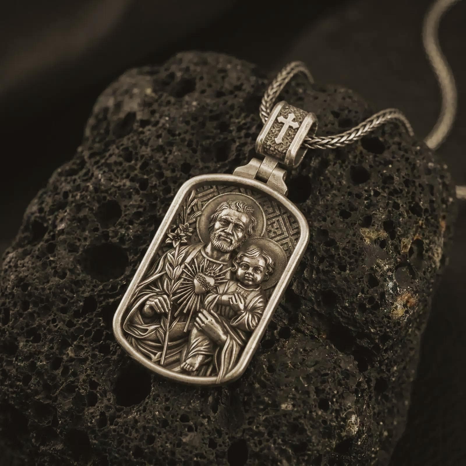 Bronze Saint Joseph and Jesus pendant necklace on textured volcanic rock, close-up studio shot highlighting the father and child relief, lily, sacred heart, and antique silver contrast.