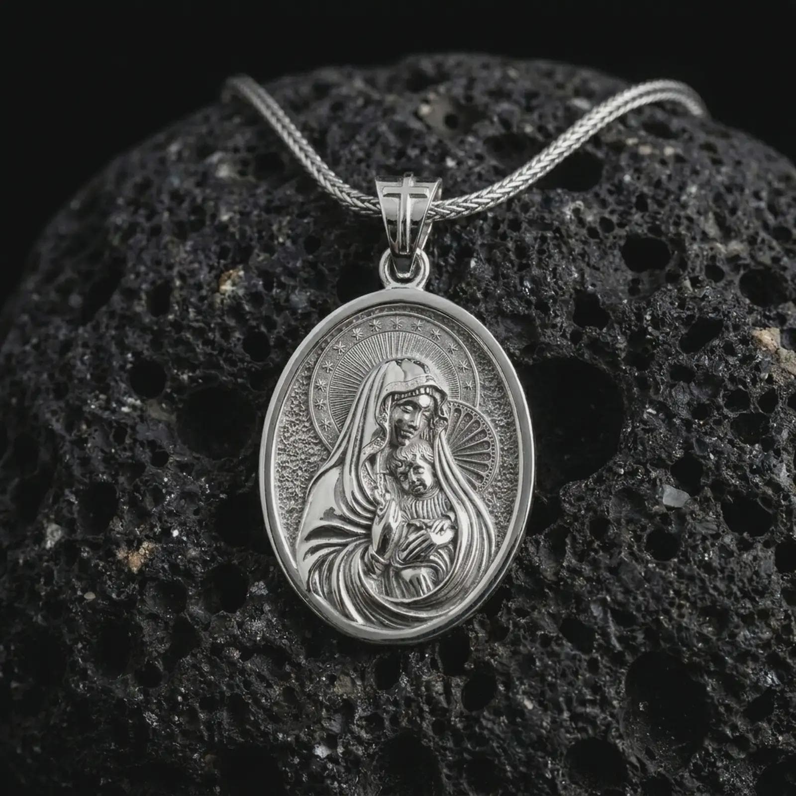 Polished silver Virgin Mary and baby Jesus pendant necklace on textured volcanic rock, centered macro jewelry photo with detailed devotional relief and bright silver highlights.