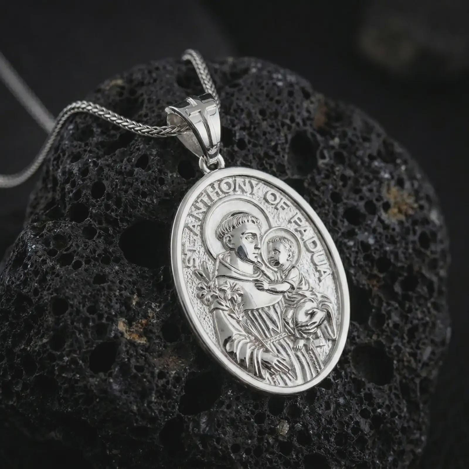 Polished silver Saint Anthony of Padua pendant necklace on textured volcanic rock, centered macro jewelry photo with detailed Catholic medal engraving and bright silver highlights.