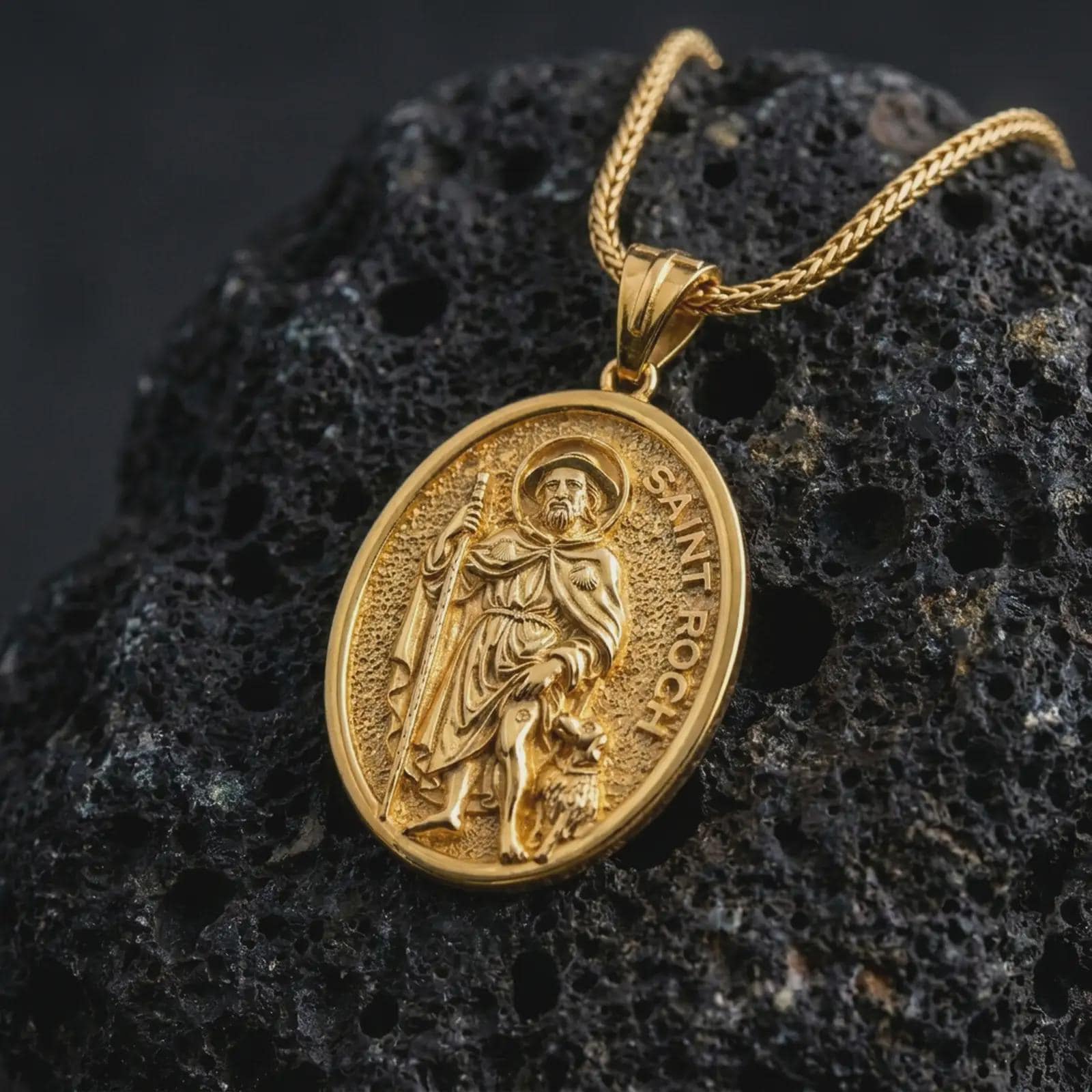 Gold plated Saint Roch pendant necklace on textured volcanic rock, premium close-up studio composition emphasizing the pilgrim saint figure, staff, and faithful dog symbolism.